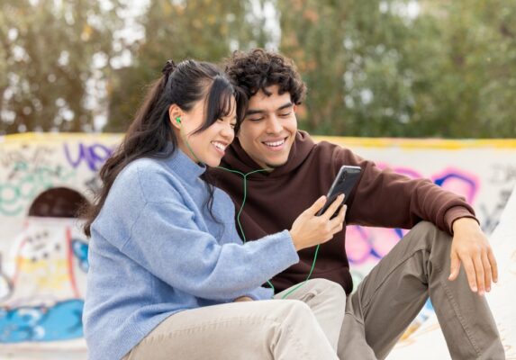Kaksi hymyilevää aikuista istuu vierekkäin skeittipuistossa. Toisella on kädessään kännykkä, jota he katsovat ja kuuntelevat jakamalla vihreät nappikuulokkeet.