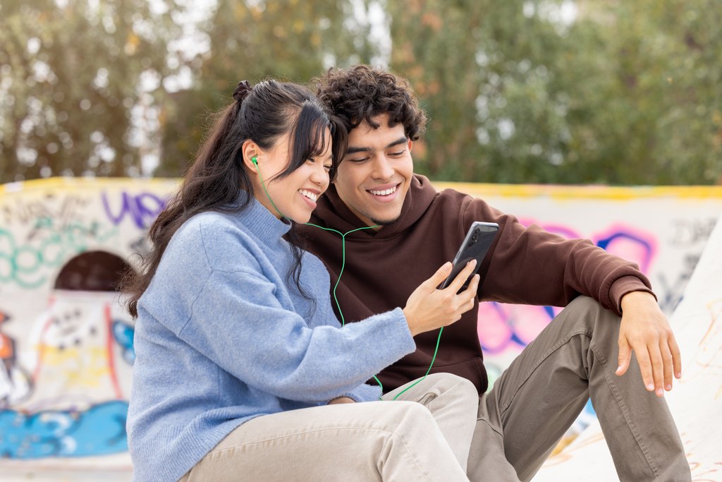 Kaksi hymyilevää aikuista istuu vierekkäin skeittipuistossa. Toisella on kädessään kännykkä, jota he katsovat ja kuuntelevat jakamalla vihreät nappikuulokkeet.